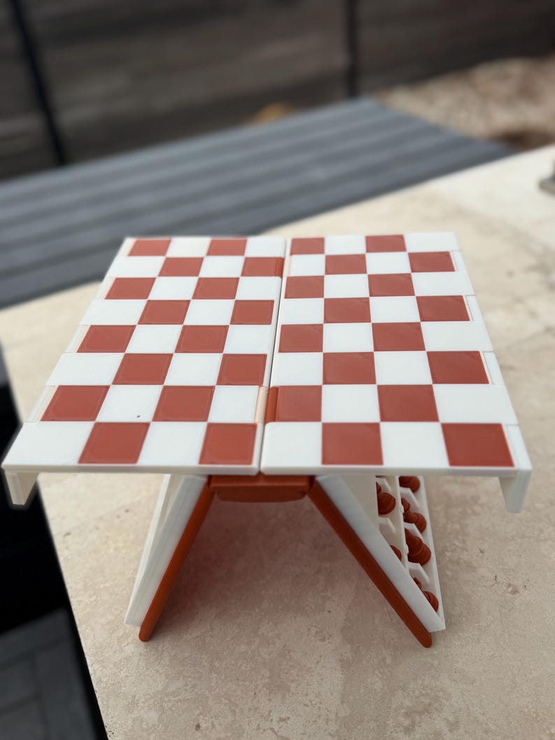 Puede incluir: Un peque&ntilde;o juego de ajedrez plegable con un tablero a cuadros rojos y blancos. El tablero est&aacute; sostenido por patas blancas y naranjas. Dise&ntilde;ado para la portabilidad, es ideal para viajes o espacios reducidos. Mide aproximadamente 25 cm x 25 cm.