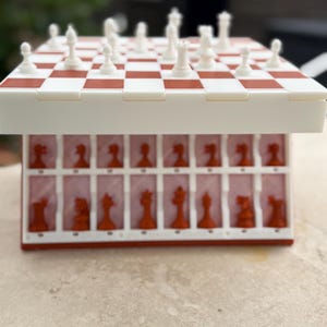 Puede incluir: Un juego de ajedrez de viaje rojo y blanco. El tablero de ajedrez es a cuadros rojos y blancos. Las piezas de ajedrez son blancas y rojas. Las piezas de ajedrez se guardan en un compartimento debajo del tablero.