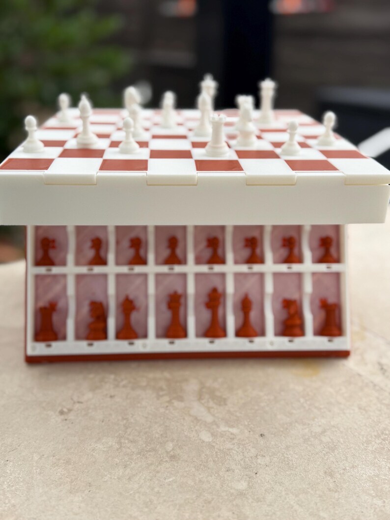 Puede incluir: Un juego de ajedrez de viaje rojo y blanco. El tablero de ajedrez es a cuadros rojos y blancos. Las piezas de ajedrez son blancas y rojas. Las piezas de ajedrez se guardan en un compartimento debajo del tablero.