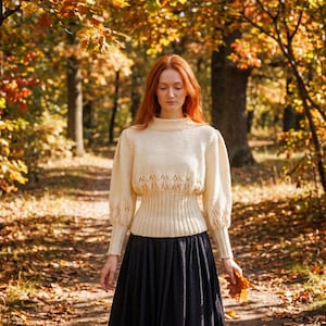 May include: A cream-colored sweater with a high neckline and long, puffed sleeves. The sweater features a decorative openwork design across the midriff and is paired with a dark blue skirt. The person is holding a fallen leaf in a forest setting.