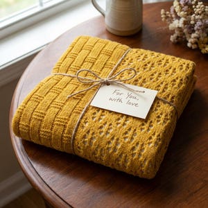 May include: A folded mustard yellow knitted blanket tied with twine and a small tag that reads "For You, with love." The blanket has a textured weave pattern on one side and a lace-like design on the other. The blanket is on a wooden table.
