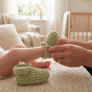 Può includere: Un paio di scarpine da neonato lavorate a maglia verde chiaro. Una viene infilata sul piedino di un bambino, l'altra poggia su una coperta di maglia color crema. Le scarpine sono realizzate con filato morbido e strutturato.