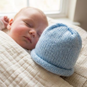 May include: A light blue knitted baby hat with a folded brim. The hat is made of soft yarn and is resting on a cream-colored blanket. The hat is approximately 6 inches in diameter.