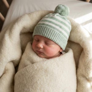 May include: A sleeping newborn baby wrapped in a cream blanket, wearing a mint green and white striped knitted hat with a pom-pom. The baby is in a crib.