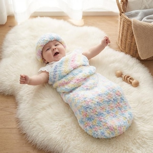 May include: A baby wearing a pastel-colored knitted hat and swaddled in a matching sleep sack. The sleep sack and hat are in shades of blue, pink, yellow, and white. The baby is lying on a white fluffy rug.