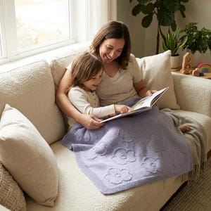 Può includere: Una coperta lavorata a maglia lilla chiaro con un motivo floreale. La coperta è drappeggiata su una persona e un bambino seduti su un divano color crema. La coperta ha un aspetto strutturato e un bordo decorativo.