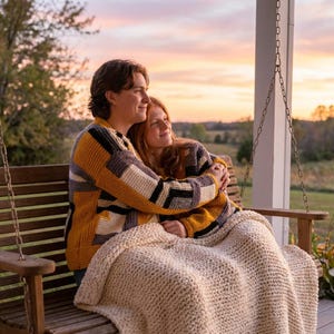 May include: Two people are seated on a wooden porch swing, embracing each other. They are wearing patchwork sweaters in shades of yellow, gray, and black. A cream-colored, chunky knit blanket covers their laps. The background features a sunset.