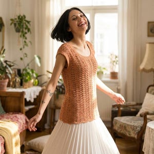 May include: A light brown, short-sleeved crochet top with a scoop neck. The top is paired with a white pleated skirt. The image is taken indoors with natural light, creating a warm and inviting atmosphere.