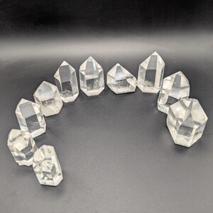 May include: A collection of clear quartz crystal points arranged in a semi-circle against a black background. Each crystal has a faceted, pointed top and a clear, translucent appearance. The crystals vary slightly in size and shape.