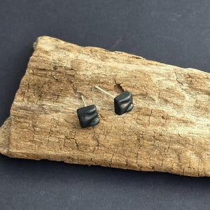 May include: A pair of small, square-shaped black stud earrings with a subtle, wavy, three-dimensional surface texture. Each earring features a thin, metallic post extending from the back, designed for secure attachment to the earlobe.