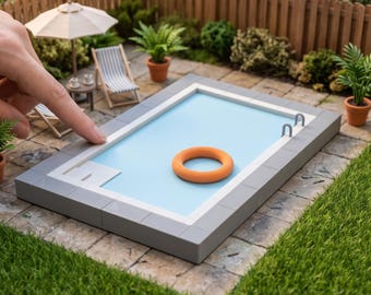 May include: A miniature rectangular swimming pool model with a light blue interior, a white border, and a single orange life ring floating on the surface. The model features two small metal-colored ladder rungs on one side and a gray tiled deck.