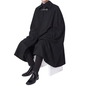 Op de afbeelding: Een zwarte cape met een gespsluiting bij de hals. De cape is lang en vloeiend, met een klassiek ontwerp. De persoon draagt een zwarte broek en zwarte leren laarzen.