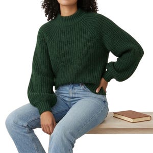 Może przedstawiać: Ciemnozielony, gruby sweter z wysokim dekoltem i długimi rękawami. Sweter jest połączony z jasnoniebieskimi dżinsami. Mała, brązowa książka spoczywa na jasnej drewnianej powierzchni.