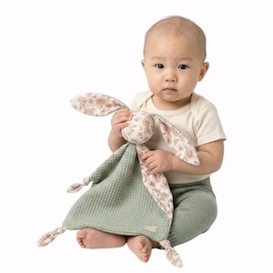 May include: A baby holds a soft, green waffle-textured security blanket with a bunny head and floral print ears. The bunny head and ears have a cream background with pink and brown floral patterns. The blanket has knotted corners.
