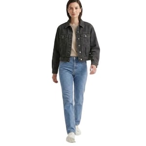 May include: A woman wearing a black denim jacket, a beige t-shirt, and light blue jeans. The jacket has button closures and pockets. The jeans are straight-legged. She is wearing white sneakers.