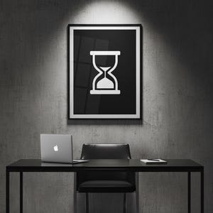 Könnte beinhalten: Ein schwarzer gerahmter Druck mit einem weißen Stundenglas-Symbol hängt über einem schwarzen Schreibtisch. Ein Laptop, ein schwarzer Stuhl und ein Notizblock befinden sich auf dem Schreibtisch. Der Hintergrund ist eine graue Betonwand.