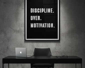 May include: A black framed print with the words "DISCIPLINE. OVER. MOTIVATION." hangs above a black desk. A laptop, chair, and notepad are on the desk. The background is a gray concrete wall.