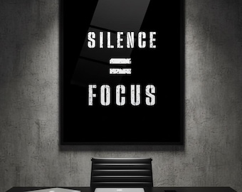 May include: A black framed poster with the words "SILENCE = FOCUS" in white, displayed above a black desk. A laptop and a smartphone are on the desk. A black chair is positioned in front of the desk. The background is a gray concrete wall.