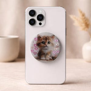 May include: A white smartphone with a pop-up grip featuring a watercolor painting of a kitten. The kitten is brown and white with pink hearts around it. The phone has a triple-lens camera.