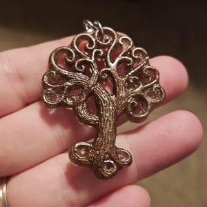 May include: A bronze-colored tree of life pendant with intricate, swirling details. The pendant features a detailed trunk and branches, with a small loop at the top for a chain. The design is symmetrical and ornate, suitable for jewelry.