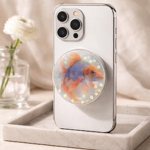 May include: A white smartphone with a decorative pop-up grip featuring a goldfish design. The fish has orange and blue coloring, with iridescent glitter accents. The phone is resting on a marble tray.