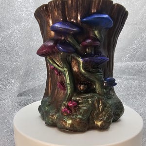 May include: A cylindrical container shaped like a tree stump with a metallic bronze finish. The exterior features sculpted mushrooms in shades of iridescent blue, purple, and pink, with green stems emerging from the base of the textured tree trunk.