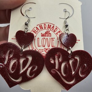 May include: Red heart-shaped earrings with the word "LOVE" cut out. The earrings have a textured surface and are attached to silver-colored fish hook ear wires. The earrings are hanging on a cream-colored card with the words "HANDMADE WITH LOVE" printed on it.