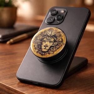 May include: A black smartphone with a gold and black pop socket featuring a detailed lion design. The pop socket has a glittery gold background and a black base, attached to the phone's back.