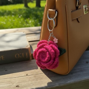 Pode incluir: Um chaveiro de rosa fúcsia feito à mão em crochê com uma pequena flor rosa e um detalhe de folha verde, preso a uma bolsa de couro castanho. A bolsa está sobre uma superfície de madeira, com um livro ao fundo.