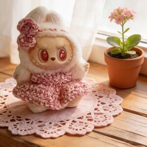 May include: A plush doll with a bunny-like hood and a pink crocheted dress. The doll has large, red eyes and is sitting on a pink lace doily. A small potted plant with pink flowers sits nearby on a wooden surface.