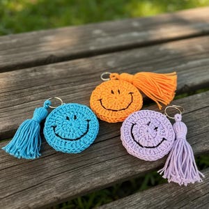 May include: Three crocheted smiley face keychains in blue, orange, and purple, each with a matching tassel. The keychains are round with a stitched smile and a gold-colored ring.