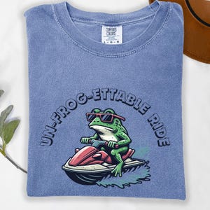 May include: A blue Comfort Colors t-shirt featuring a cartoon frog wearing sunglasses and riding a jet ski. The text "UN-FROG-ETTABLE RIDE" is arched around the frog. The shirt is folded and has a tag in the upper center.