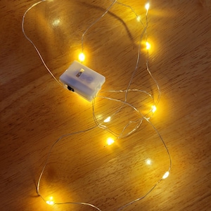 Op de afbeelding: Een reeks warmwitte LED-feeërieke lampjes met een kleine witte batterijhouder. De lampjes zitten op een dunne zilverkleurige draad en de batterijhouder heeft een aan/uit-schakelaar. De lampjes liggen verspreid op een houten oppervlak.