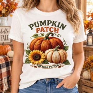 Pode incluir: Uma t-shirt branca com um gráfico de duas abóboras com textura de malha e um girassol grande. O texto PUMPKIN PATCH está impresso em verde escuro e laranja acima das abóboras, e as palavras FRESHLY PICKED aparecem numa faixa abaixo.