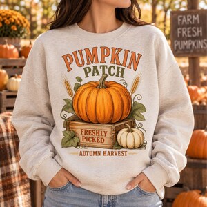 Pode incluir: Uma camisola de gola redonda em cinzento mesclado claro com um gráfico de uma abóbora laranja grande sobre uma caixa de madeira com o texto PUMPKIN PATCH, FRESHLY PICKED e AUTUMN HARVEST. Uma abóbora branca mais pequena e espigas estão presentes.