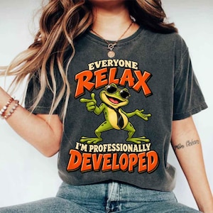 May include: A charcoal gray short-sleeve t-shirt featuring a graphic of a green frog wearing sunglasses and a black necktie. The text reads "EVERYONE RELAX I'M PROFESSIONALLY DEVELOPED" in bold orange lettering above and below the frog illustration.