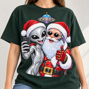 Pode incluir: Uma t-shirt verde-escura com um gráfico de um alienígena cinzento e o Pai Natal a usar gorros vermelhos e brancos. O alienígena segura um smartphone preto para tirar uma selfie, enquanto o Pai Natal faz um gesto de positivo.