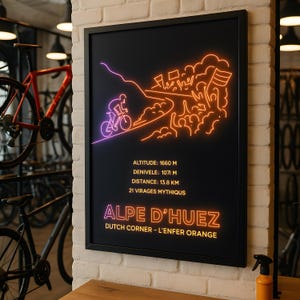 Peut inclure: Affiche encadrée noire avec une illustration de style néon d'un cycliste sur une route de montagne. L'affiche comprend le texte "ALPE D'HUEZ" et les détails : altitude 1660 m, distance 13,8 km et 21 virages. La phrase "DUTCH CORNER - L'ENFER ORANGE" est également incluse.