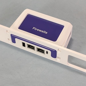 May include: A white rack-mount bracket holding a Firewalla network security device. The device features a purple top panel with the white text Firewalla, and the front panel includes two Ethernet ports, a USB port, and a small circular port.