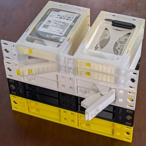 May include: A stack of four 3D-printed hard drive caddy adapters in translucent white, solid white, black, and yellow. The top two units contain Seagate hard drives with visible SATA connectors. Each caddy features a front-facing latch mechanism.