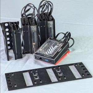 Może przedstawiać: Zestaw czarnych komponentów komputerowych, w tym kilka czarnych obudów z dołączonymi kablami, urządzenie z przezroczystym wierzchem odsłaniającym wewnętrzne obwody oraz czarny wspornik montażowy. Urządzenie ma napis "X&A".