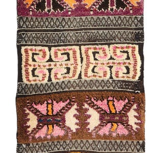 Peut inclure: Une tenture murale textile tissée rectangulaire présentant un fond brun foncé avec des bandes horizontales de motifs géométriques et abstraits en relief dans des tons orange, rose, violet, crème et noir, finie avec un bord frangé.