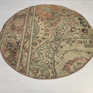 May include: A round area rug featuring a distressed, multi-tonal pattern in shades of muted orange, beige, and dark brown. The design incorporates intricate floral and geometric motifs with a visible weathered texture and a stitched circular border.