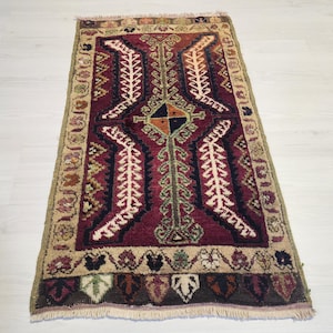 May include: A rectangular area rug featuring a deep burgundy central field with a dark blue geometric motif, white leaf-like patterns, and a central diamond divided into four colored sections. The border is beige with floral accents.