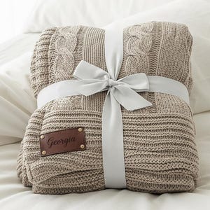 May include: A folded beige cable knit blanket tied with a silver ribbon. The blanket has a leather patch that reads "Georgia". The blanket is on a white bed.