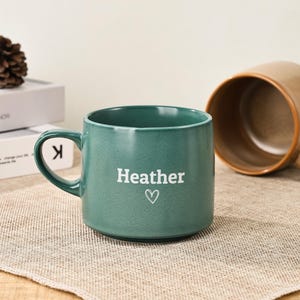 May include: A teal ceramic mug with the name "Heather" in white, accompanied by a small heart symbol. The mug has a rounded handle and a speckled texture. Another brown mug is visible in the background.