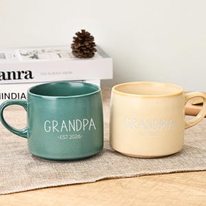 Peut inclure: Deux mugs en céramique, un vert d'eau et un crème, avec des anses. Le mug vert d'eau porte l'inscription "GRANDPA - EST 2026" en lettres blanches. Le mug crème porte l'inscription "GRANDMA - EST 2026" en lettres blanches.