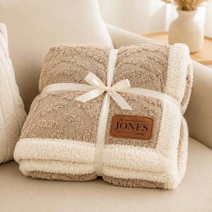 May include: A folded tan throw blanket with a textured leaf pattern and a white sherpa fleece border. The blanket is tied with a cream-coloured ribbon and features a brown leather-like patch with the text "The JONES Family" printed in black.