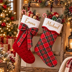 May include: Two Christmas stockings hanging on a wooden ladder. The left stocking is quilted red fabric with a white cuff embroidered with the name Emma and a plaid bow. The right stocking is red and green plaid with a white cuff embroidered with Michael.