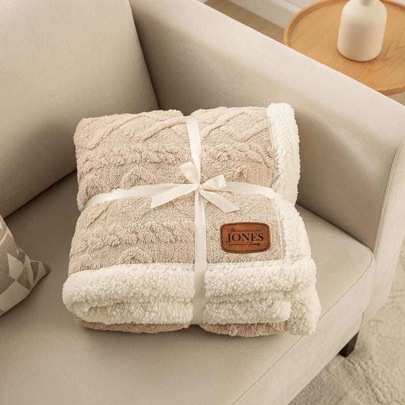 May include: A folded beige throw blanket with a textured cable knit pattern on one side and a white sherpa fleece lining on the other. A white ribbon is tied around the centre, and a brown leather patch features the text The JONES Family.